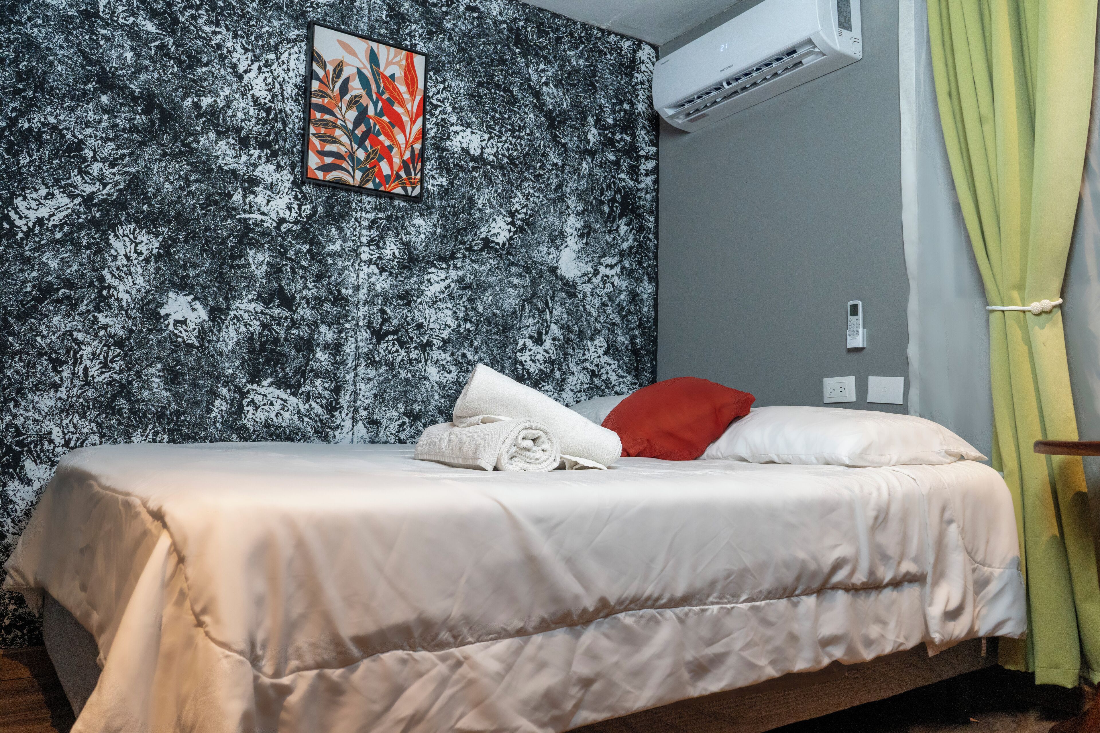 A neatly made bed with white linens and a red pillow, a wall-mounted air conditioner, and a painting above the bed.