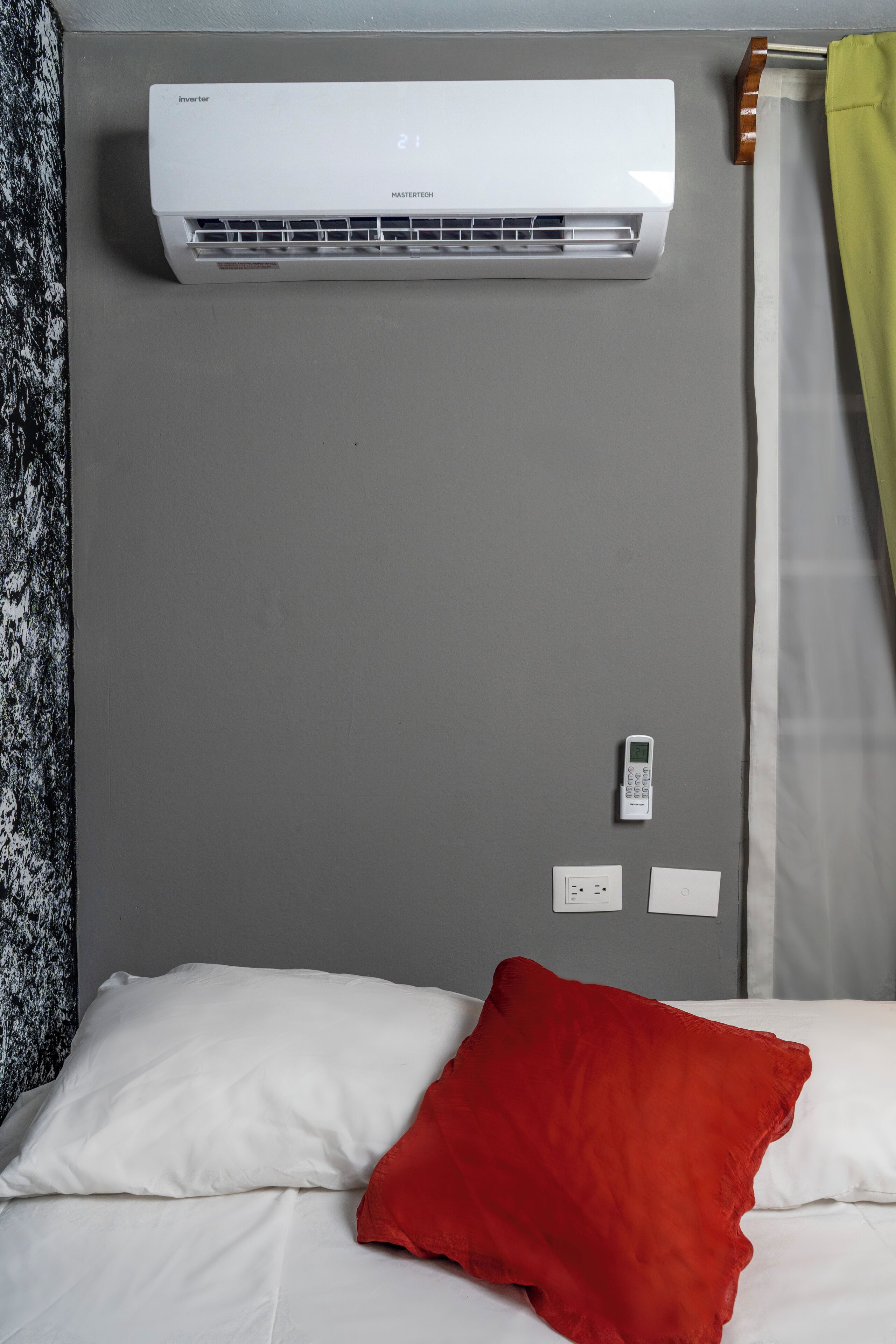 Une pièce avec un lit, un oreiller rouge, un climatiseur blanc et un panneau de commande fixé au mur.