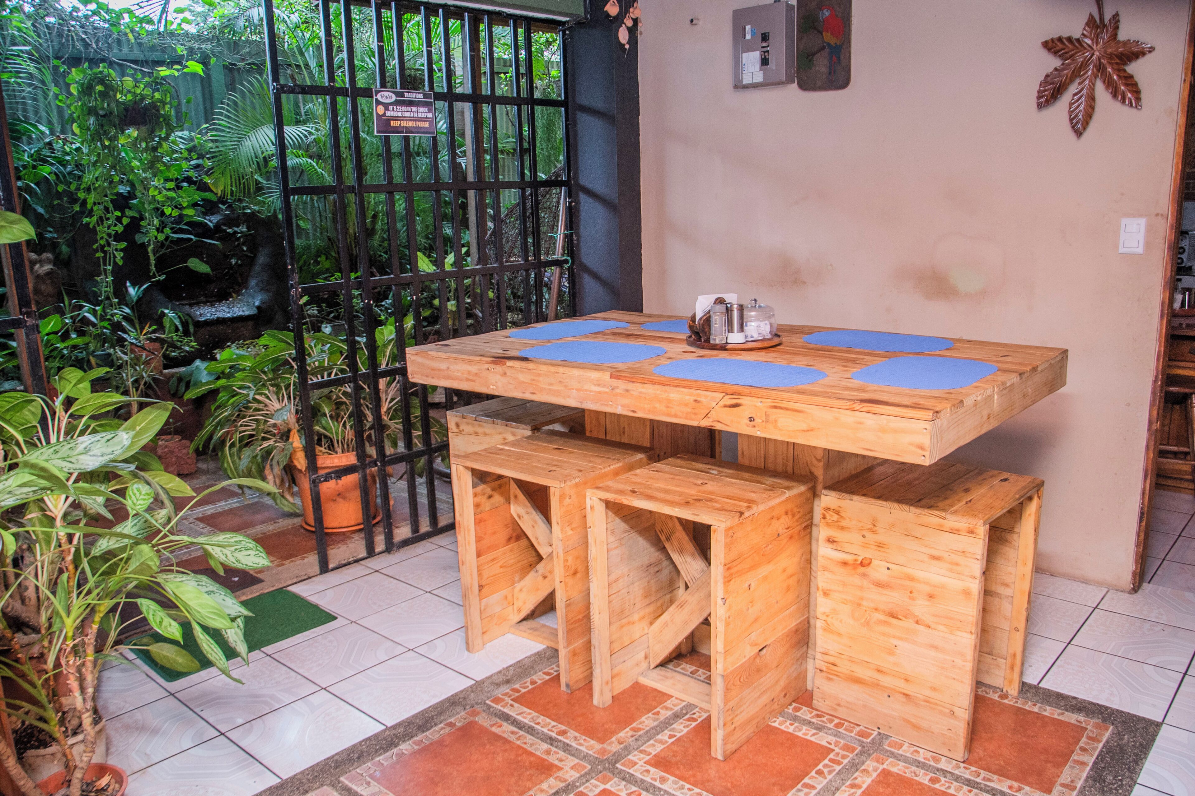 Une table en bois, agrémentée de sets de table bleus et entourée de bancs, avec des plantes en pot et un portail en métal noir.