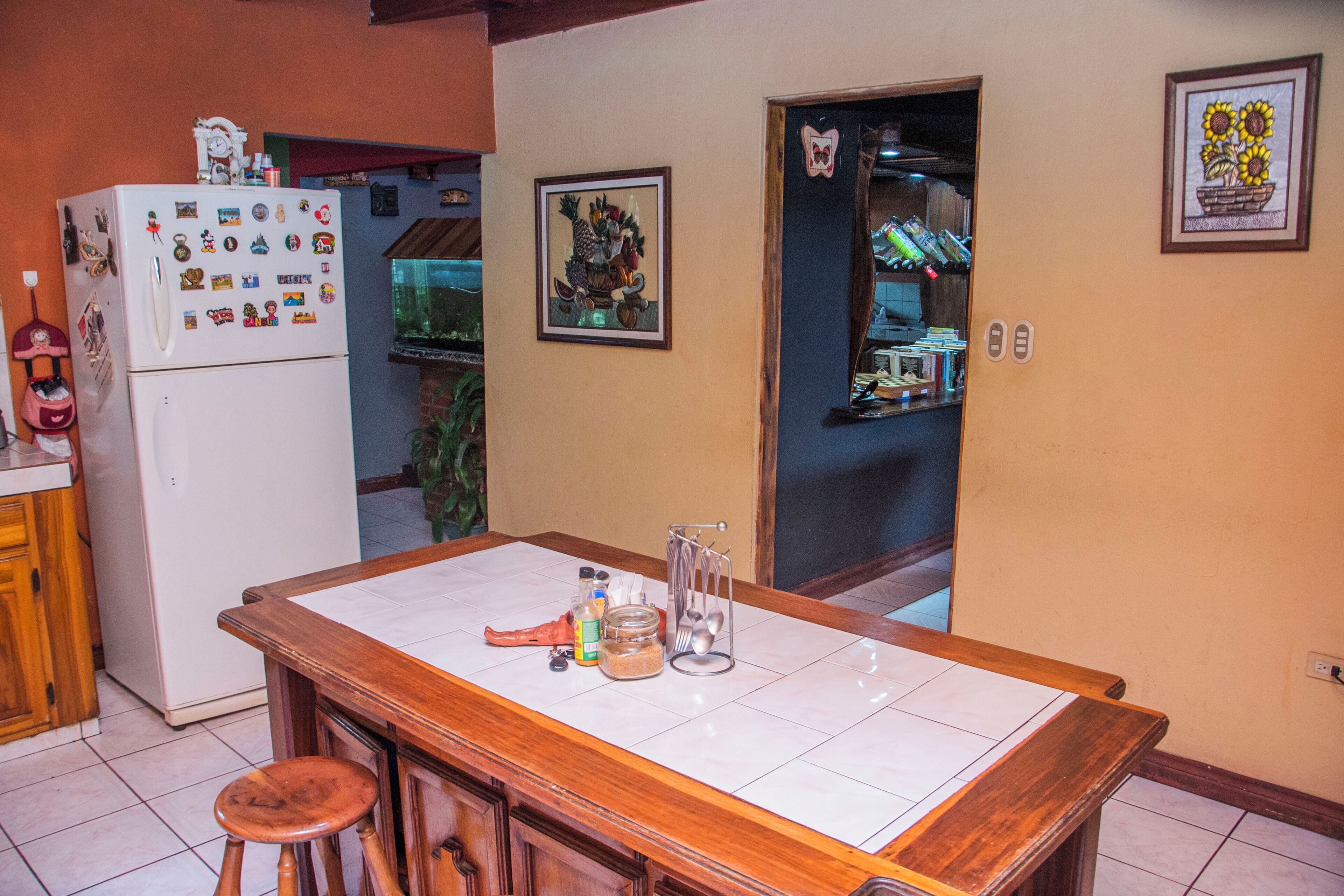 A kitchen with a wooden bar counter, a refrigerator with magnets, a framed picture on the wall, and a doorway leading to another room.
