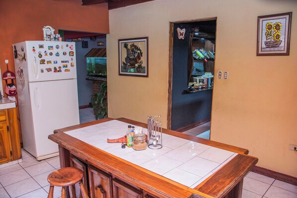 Una cocina con barra de madera, un refrigerador con imanes, un cuadro en la pared y un acceso a otra habitación.