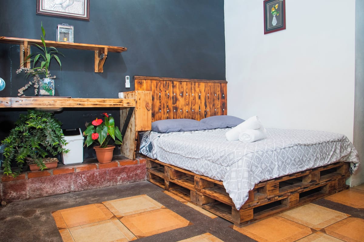 A rustic bedroom with a wooden bed, a small table with plants, and a dark wall with framed pictures.