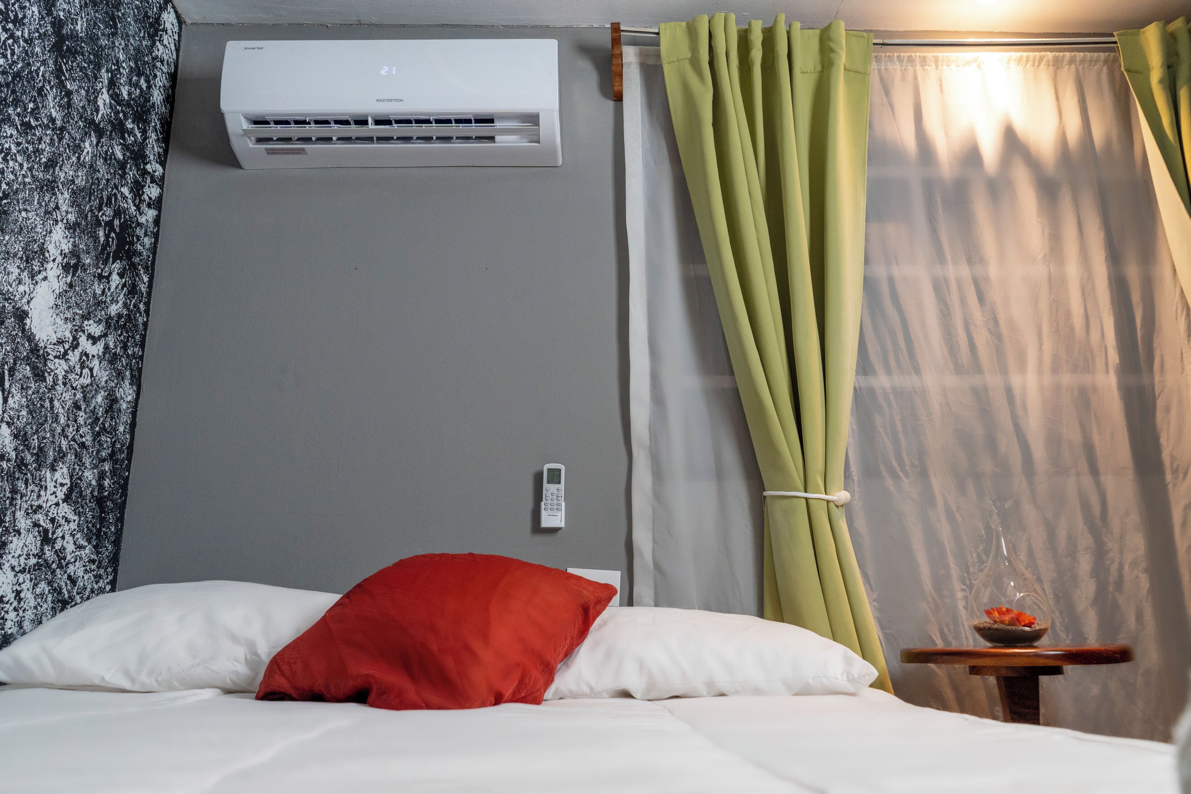 A hotel room with a bed, a wall-mounted air conditioner, a bedside table with a bowl of fruit, and curtains.