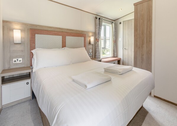 A neatly made bed with white linens and towels, a bedside table with a lamp, and a window with curtains.
