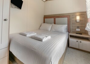 Cama bem arrumada com lençóis e toalhas brancos, cabeceira de madeira, mesa de cabeceira com controle remoto e televisão na parede.