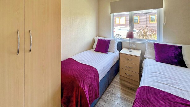 A bedroom with two beds, a wooden wardrobe, a window with blinds, and a bedside table with a lamp.
