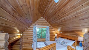 Quarto de cabana de toras com uma cama, uma janela com cortinas, uma mesa de cabeceira e um abajur.