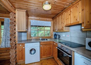 A wooden cabin kitchen with a washing machine, oven, and a window with a view of greenery.