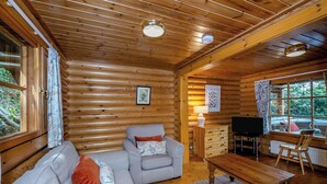 Interior de cabana de tronco com sofá, mesa de madeira, televisão e janela com vista para o exterior.