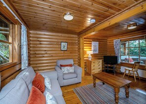 A log cabin interior with a sofa, a wooden table, a television, and a window with a view of the outdoors. - Timber lodge set in an elevated position with views of the lake. (Webbery, Bideford)