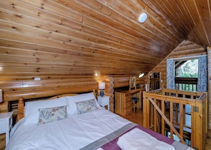 A wooden cabin bedroom with a bed, bedside table, lamp, and a view of the outdoors through a window.