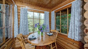 Interior de uma cabana de madeira com mesa redonda para refeições, cadeiras e janela com cortinas.