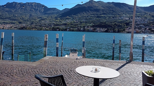 Een terras aan het water met een tafel en stoelen, houten palen en uitzicht op een meer met bergen op de achtergrond.