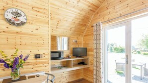 Ein Interieur einer Holzhütte mit Esstisch, Stühlen, einer Uhr, einem Fernseher und einem Fenster mit Vorhängen.