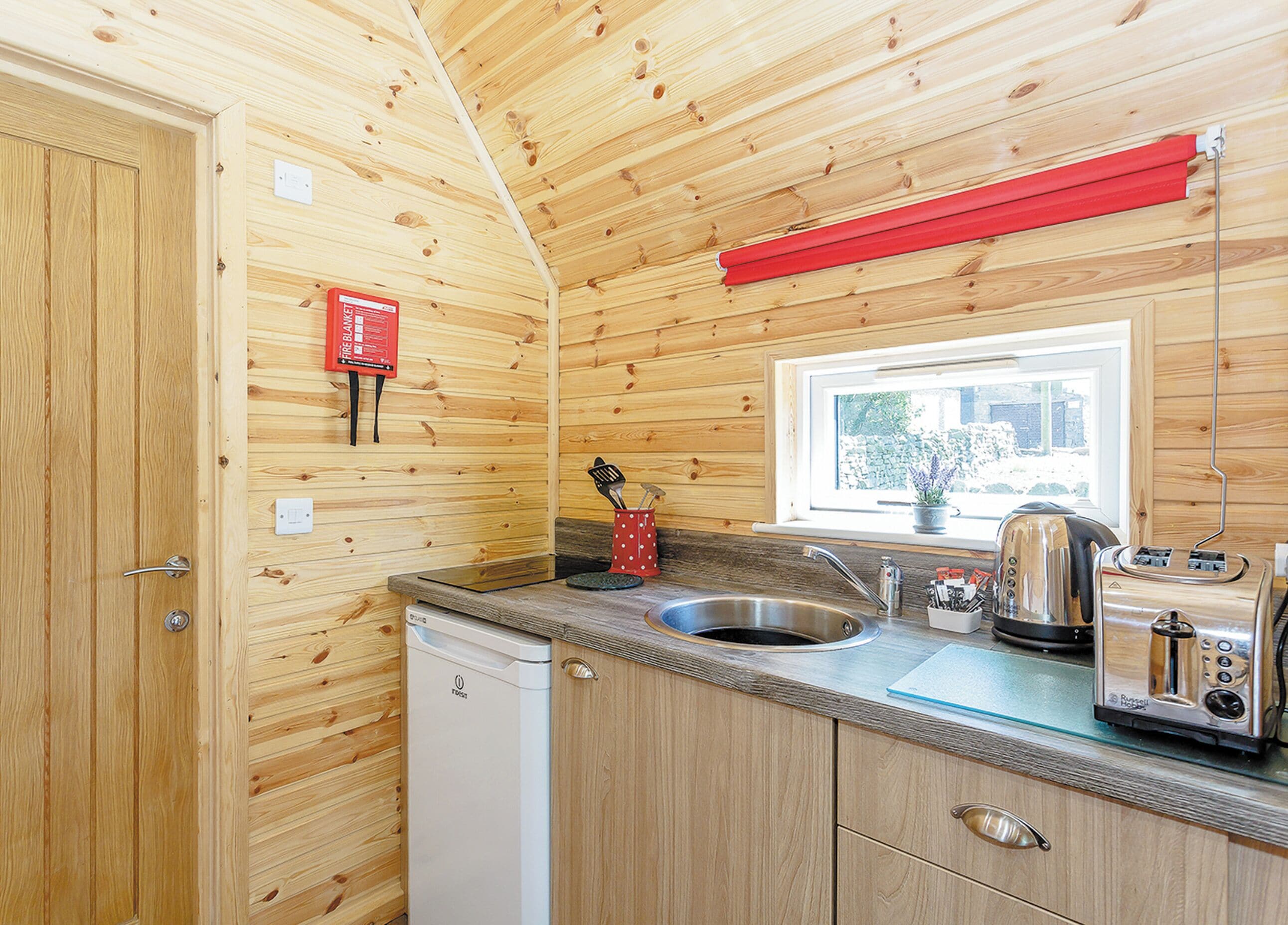 A compact kitchen with wooden walls, a stainless steel sink, a toaster, and a window with a view of the outside.