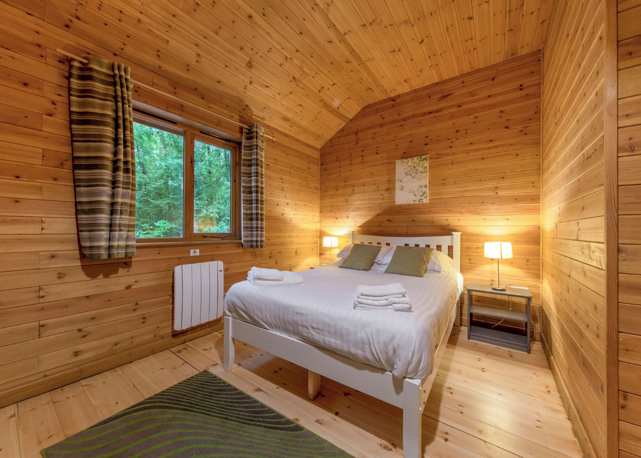 Ein Schlafzimmer einer Holzhütte mit einem Bett, zwei Nachttischlampen, einer Heizung und einem Fenster mit Vorhängen.