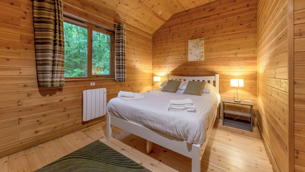 Ett sovrum i en stuga med träpanel, en säng, två sänglampor, ett element och ett fönster med gardiner.