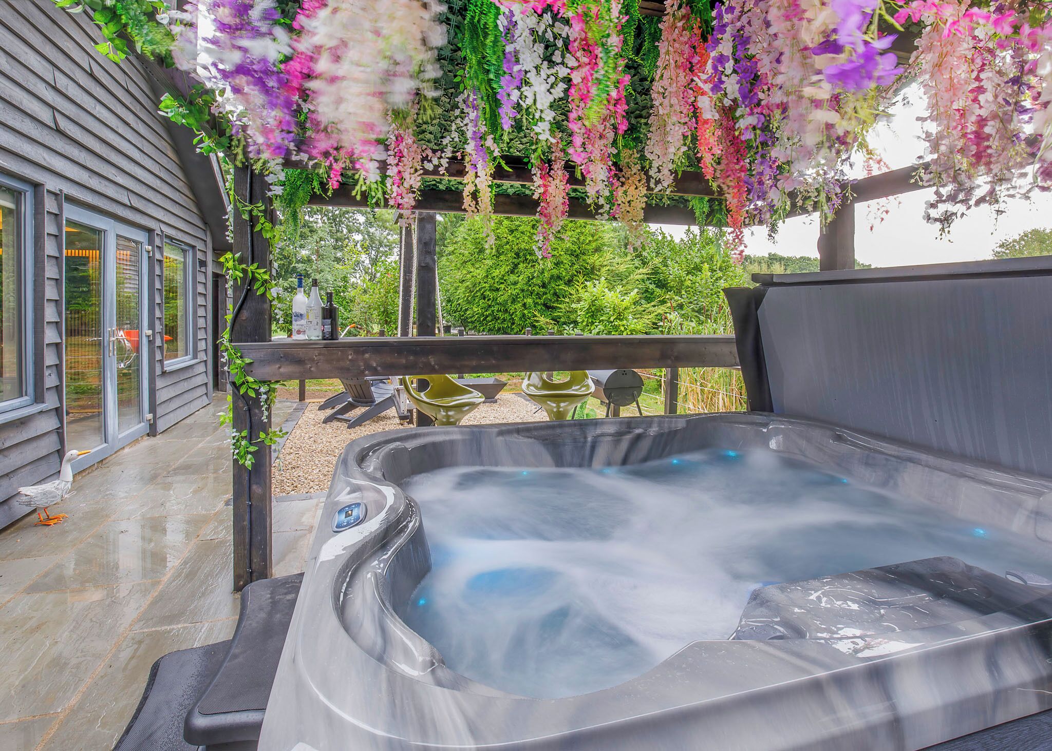 A hot tub with a wooden deck, surrounded by hanging flowers, and a view of a house in the background.