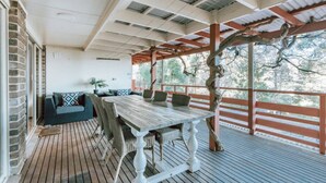 Une terrasse extérieure avec une table et des chaises en bois, un banc rembourré et une pergola soutenue par un tronc d’arbre.