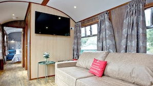 A cozy interior with a sofa, a small table, a TV mounted on the wall, and a window with curtains. - Brunel Boutique Railway Carriage 1 (Dawlish Warren)