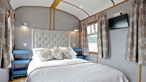 A bedroom with a tufted headboard, a bed with a white quilt, a blue bedside table, and a television mounted on the wall. - Brunel Boutique Railway Carriage 1 (Dawlish Warren)