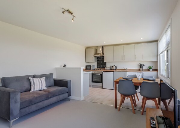 Une cuisine moderne avec des meubles blancs, un coin repas comprenant une table et des chaises, et un canapé gris agrémenté d’un coussin à rayures.