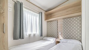 A room with two single beds, a window with curtains, patterned wallpaper, and a wooden headboard.