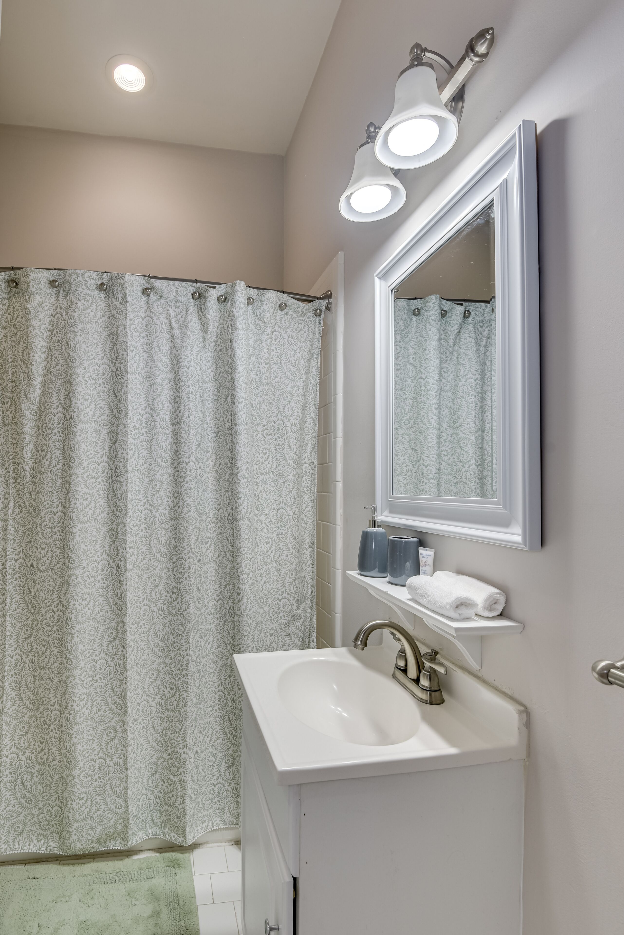 A bathroom with a shower curtain, a sink, a mirror, and a wall-mounted light fixture.