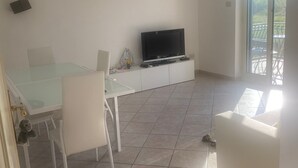 Un vaste espace de vie comprenant une table à manger, des chaises blanches, un téléviseur à écran plat posé sur un meuble blanc, et une grande porte-fenêtre coulissante donnant sur un patio extérieur.