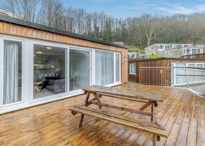 A wooden deck with a picnic table, large sliding glass doors, and a view of a forested area. - Sea Shore apartments are perfect for families. (Millendreath, Looe)