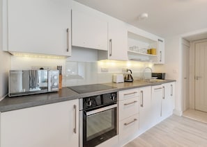 A modern kitchen with white cabinetry, a built-in oven, a toaster, and a kettle on the countertop. - Sea Shore apartments are perfect for families. (Millendreath, Looe)