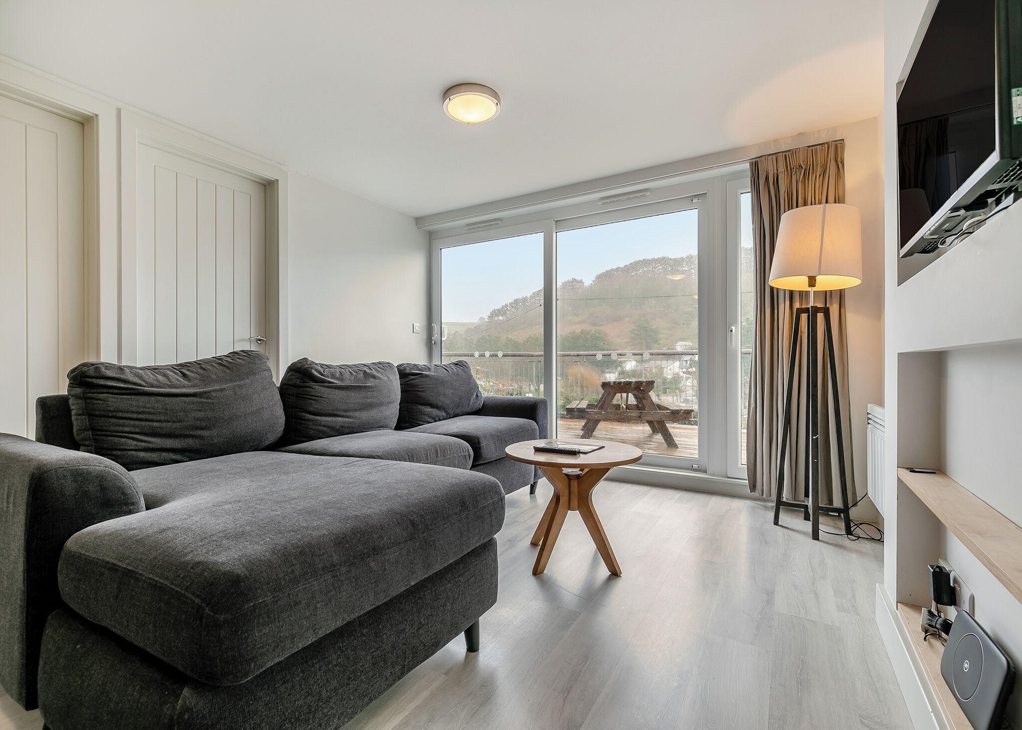 A modern living room with a grey sectional sofa, a small wooden coffee table, and a large sliding glass door leading to an outdoor area with a table and chairs.
