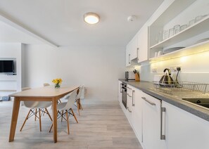 A modern kitchen with white cabinets, a wooden dining table, and a built-in oven. - Sea Shore apartments are perfect for families. (Millendreath, Looe)