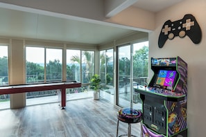 A room with a pool table, a large window with a view of greenery, and a Turtles arcade machine.