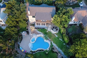 An aerial view of a house with a swimming pool, surrounded by trees and a well-maintained lawn.