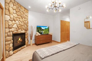 A bedroom with a stone wall fireplace, a TV on a wooden dresser, a bed with a patterned blanket, and a chandelier. - EVL Chalet at Holiday Valley (Ellicottville)