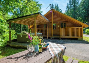 Eine Holzhütte mit einer Veranda, einem Picknicktisch mit Weingläsern und einer Flasche, und einer Badewanne im Hintergrund.