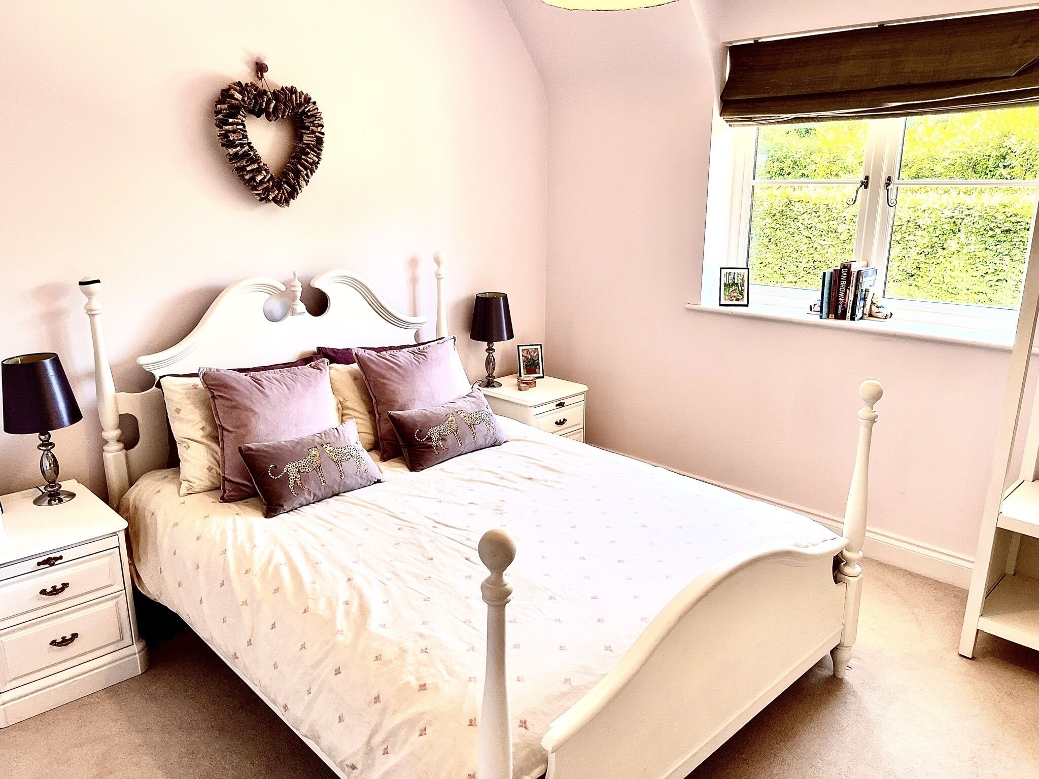 A bedroom with a white bed, multiple pillows, a nightstand with a lamp, a dresser, and a window with a view of greenery.