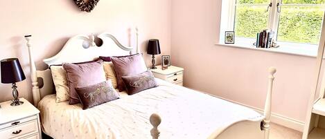 A bedroom with a white bed, multiple pillows, a nightstand with a lamp, a dresser, and a window with a view of greenery.