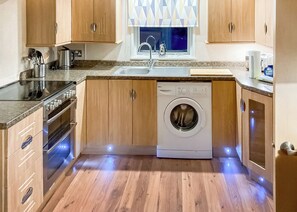 A compact kitchen with wooden cabinets, a built-in washing machine, stainless steel appliances, and a window with patterned blinds.