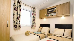 Une chambre avec deux lits, une fenêtre ornée de rideaux à motifs floraux, une armoire en bois avec des étagères et un ours en peluche sur l’un des lits.