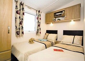 Una habitación con dos camas, una ventana con cortinas florales, un armario de madera con estantes y un osito de peluche en una de las camas.