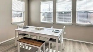 Un coin repas avec une table et un banc blancs, trois fenêtres équipées de stores et un mur gris.