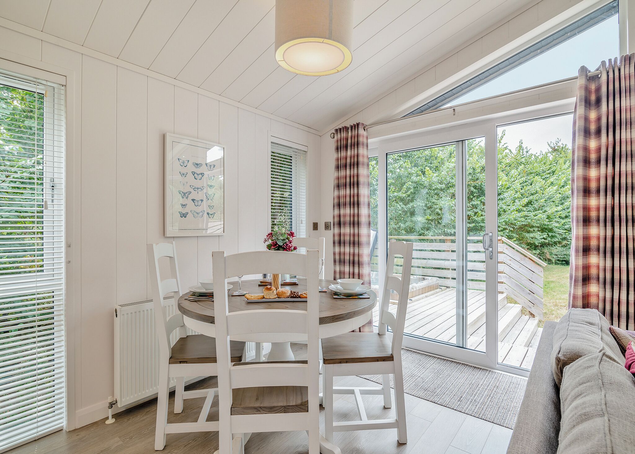 A dining area with a round table, white chairs, a flower vase, and a view of a deck through large windows.
