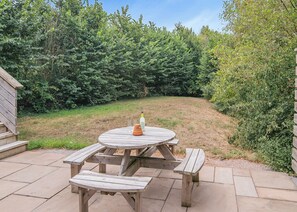 A wooden picnic table with benches, a bottle, and a candle on it, surrounded by a grassy area and dense foliage.