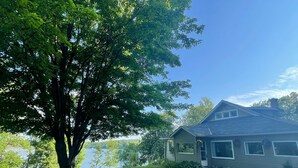 A house with a large tree in the front yard, a pathway leading to the house, and a view of a lake. - Deer Hill Cottage on 5 Lake Chain (Haliburton)