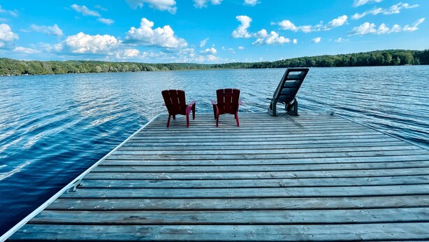 A wooden dock extending into a calm lake with two red chairs and a black lounge chair. - Deer Hill Cottage on 5 Lake Chain (Haliburton)