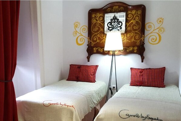 A room with two beds, each with a red plaid pillow and a white bedspread. There is a decorative headboard with a black and gold design above the beds. A floor lamp is positioned between the beds.