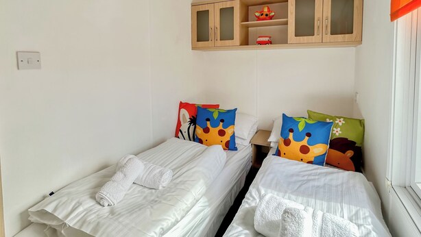 Zwei Einzelbetten mit weißer Bettwäsche und bunten Kissen, ein kleiner Holzschrank darüber und ein Fenster mit Jalousien.