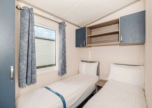 Una habitación compacta con dos camas, una ventana con cortinas, un pequeño estante de madera y un enchufe.